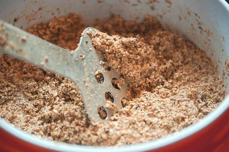 A close-up of a metal masher mixing a coarse, brown mixture in a red container.