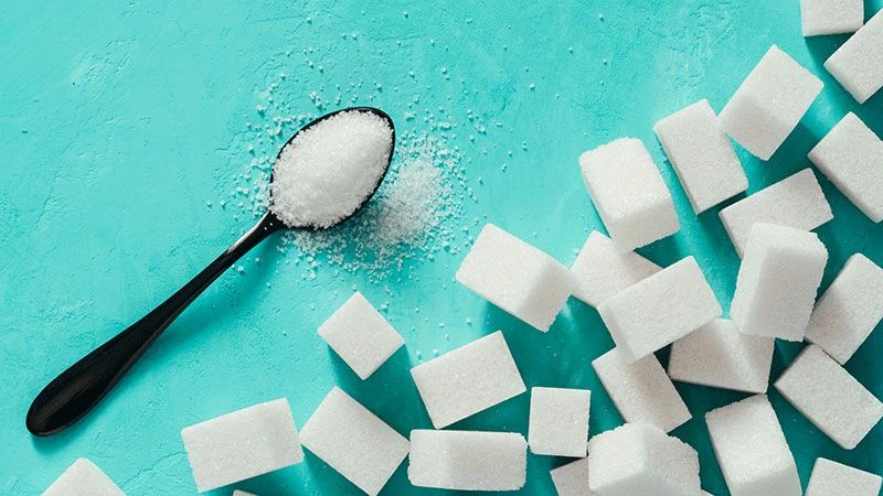 Black spoon with granulated sugar and sugar cubes on turquoise surface.